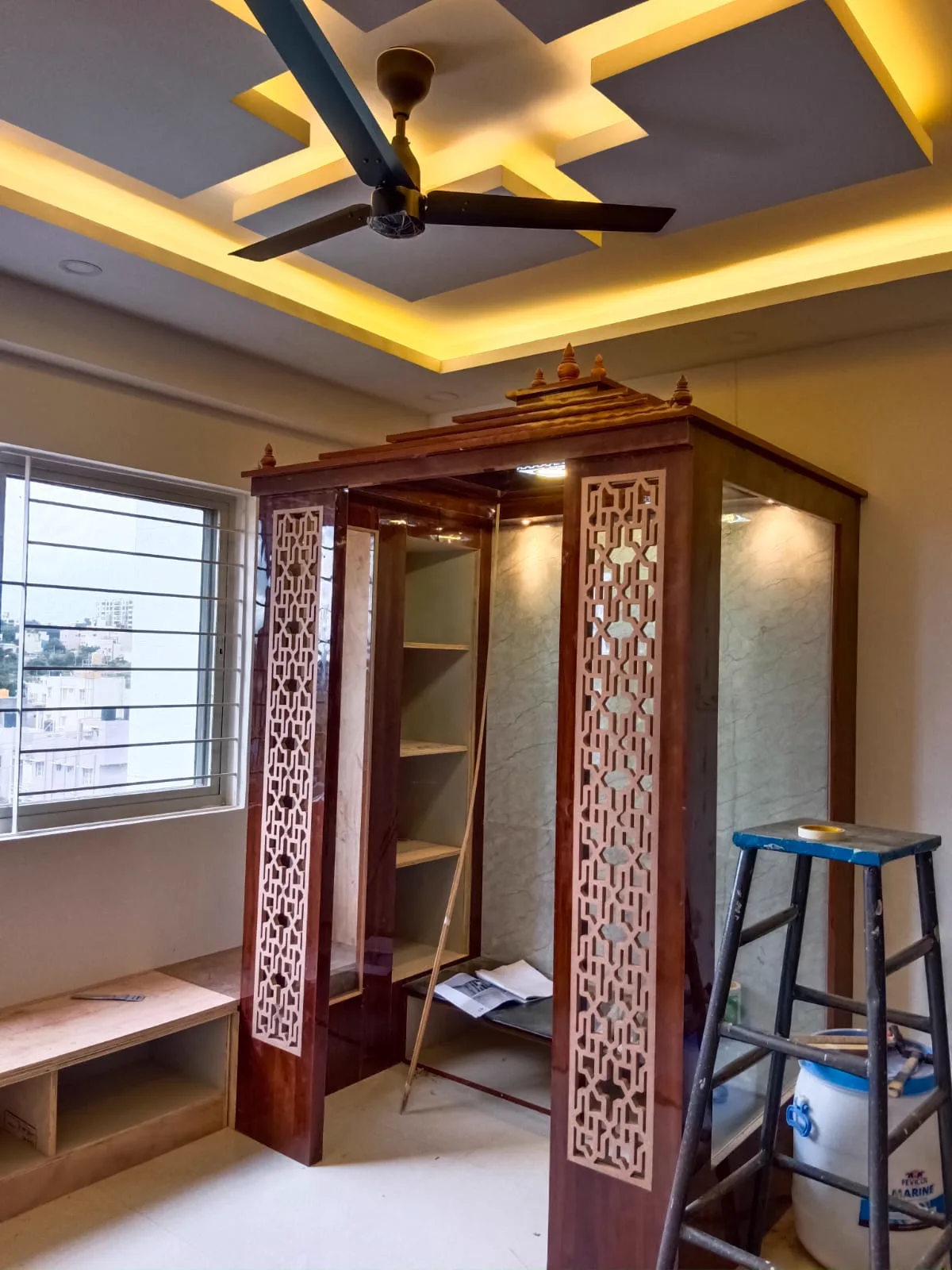 Wooden pooja unit with carved jaali screens and a lit false ceiling